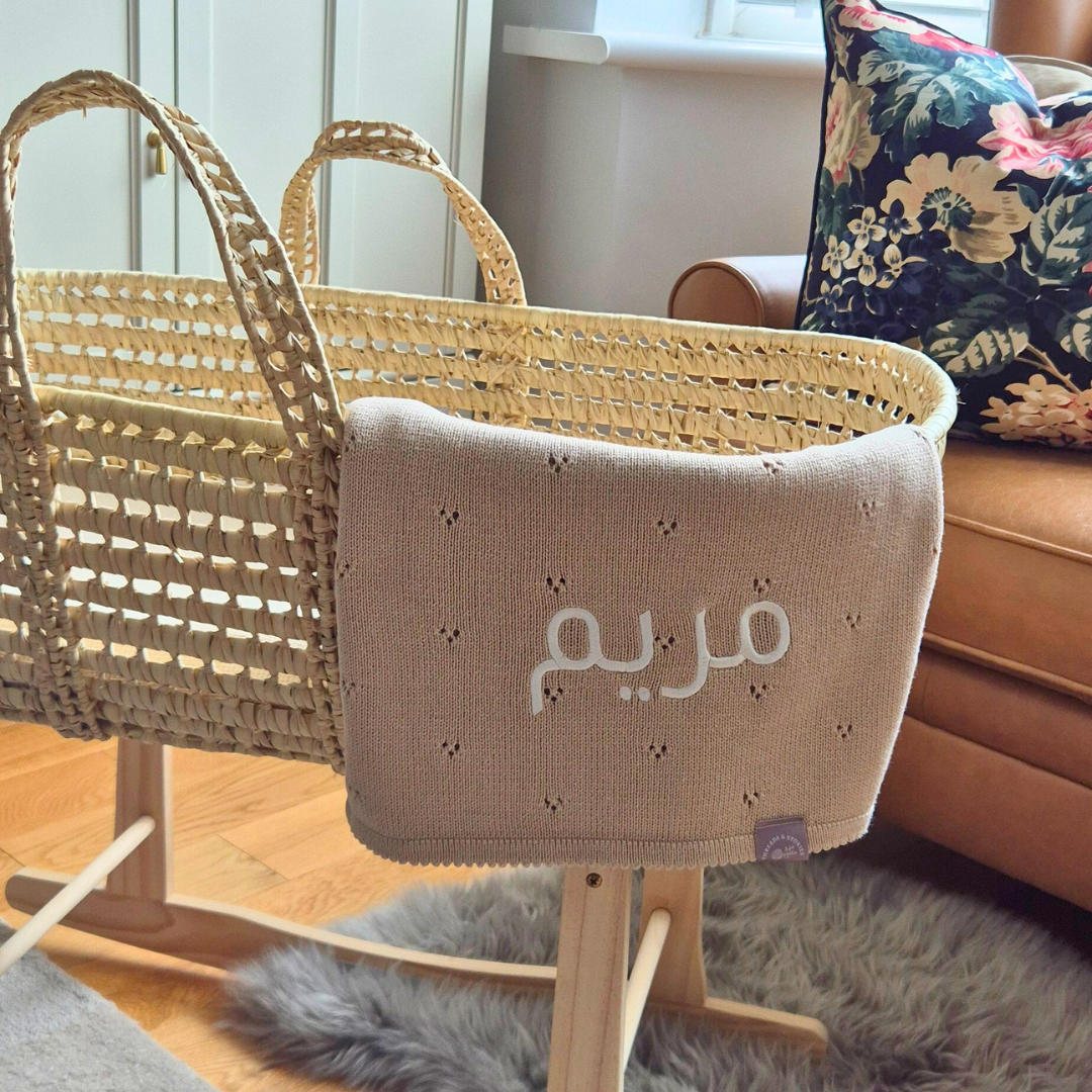 Wicker baby crib with a beige blanket featuring Arabic text on a wooden floor.