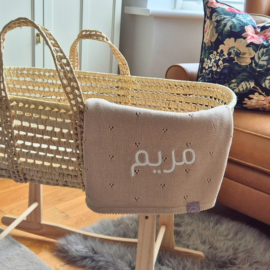 Wicker baby crib with a beige blanket featuring Arabic text on a wooden floor.